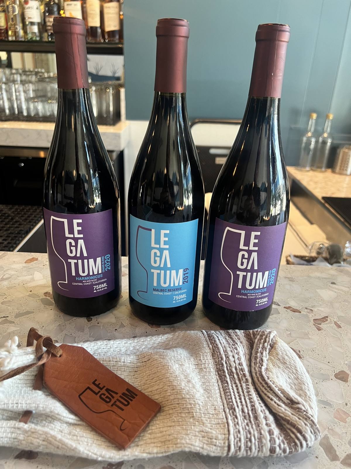 Two purple-label 2020 Harmonicus bottles flanking a blue-label 2019 Malbec Reserve with a woven sleeve and leather tag in front