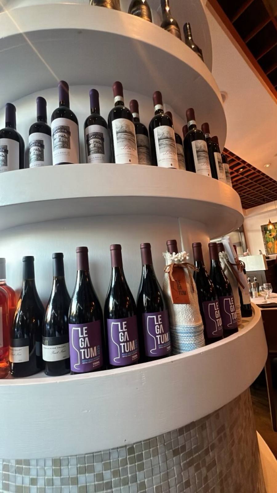 Legatum wines on Toscanova’s curved wine shelf, including the purple-label 2020 Petite Sirah Reserve beside a bottle in its hand-woven sleeve and leather tag